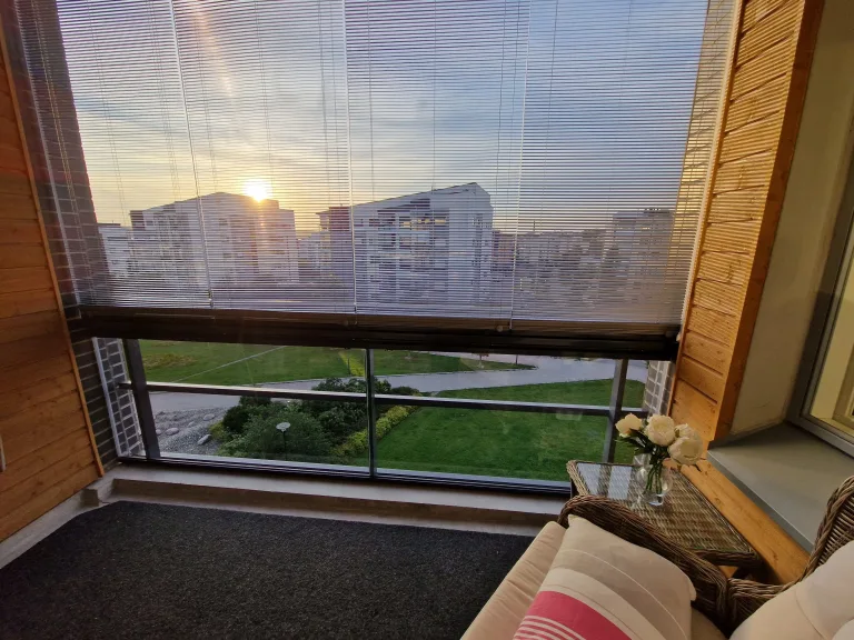 Parveke, jossa iso lasi. Auringonsäteet heijastavat kaukaisten kerrostalojen takaa. Parvekekaihtimet ovat puoliauki.