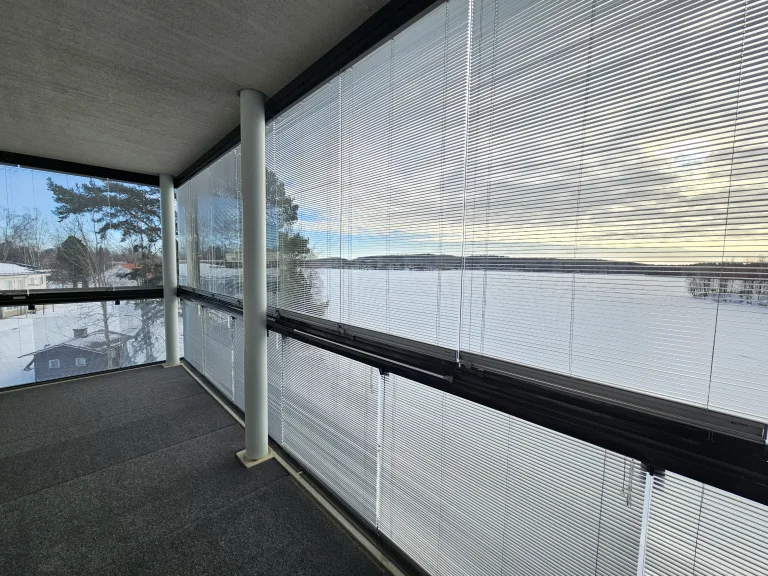 Iso Parveke, jossa kokopitkä iso lasi. Ulkona talvi ja lunta. Lähellä siintää järvi. Parvekekaihtimet ovat auki ylälaseissa ja alalaseissa kiinni.